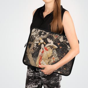 Peut inclure: Une housse d'ordinateur portable noire avec une illustration colorée d'une femme en kimono japonais traditionnel tenant une fleur. L'arrière-plan présente un arbre avec des fleurs blanches et un ciel bleu foncé.