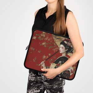 Peut inclure: Une housse d'ordinateur portable noire avec un motif japonais coloré représentant une femme tenant un parapluie. Le motif comprend des couleurs rouge, or et noir avec des motifs floraux.