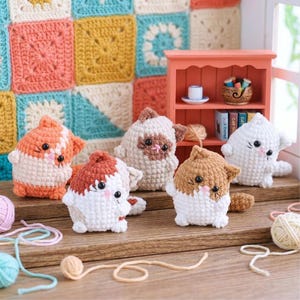 May include: A collection of five crocheted cat figurines in various colors, including orange, brown, and white. Each cat has a unique design, with black eyes and pink cheeks. The cats are displayed on a wooden surface with yarn and a miniature shelf in the background.
