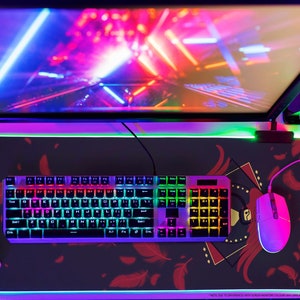 Op de afbeelding: Een zwarte gaming muismat met een kleurrijk verenontwerp. Op de muismat liggen een regenboogkleurig toetsenbord, een witte gamecontroller en een witte gamingmuis.