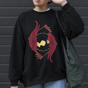 May include: Black sweatshirt with a red and yellow graphic design featuring a stylised bird with wings spread. The design is set within a diamond shape with a gold outline.