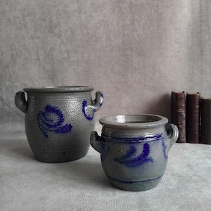 Op de afbeelding: Twee grijze stenen potten met blauwe accenten en handvatten aan de zijkanten. De grotere pot heeft een gebogen blauw ontwerp, terwijl de kleinere pot een blauwe band rond de rand en een blauw patroon op het lichaam heeft. Beide potten zijn wijd.