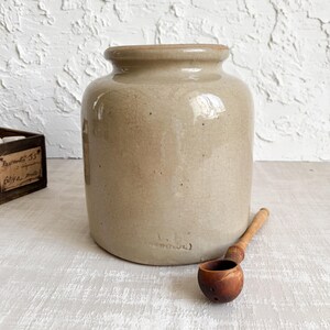 Op de afbeelding: Een cilindervormige beige stenen pot met een glanzend glazuur en zichtbare haarscheurtjes. De basis is voorzien van een reliëfstempel met de tekst "L.B. ARGENTEUIL". Een kleine, donkere houten lepel met een ronde kom ligt ernaast.