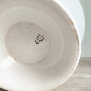 May include: A white ceramic vessel base featuring a black stamped maker's mark on the interior bottom. The mark displays the text PEXONNE inside a shield shape, accompanied by the letters FF and two small stars, indicating the origin of the piece.
