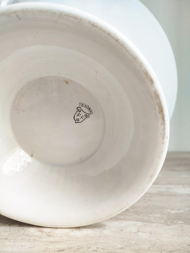 May include: A white ceramic vessel base featuring a black stamped maker's mark on the interior bottom. The mark displays the text PEXONNE inside a shield shape, accompanied by the letters FF and two small stars, indicating the origin of the piece.