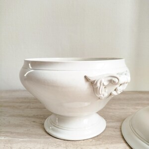 May include: A white ceramic tureen with a smooth, glossy finish, featuring a wide, rounded bowl, a pedestal base, and ornate, scrolled handles on the sides. A matching lid is positioned partially out of frame to the right of the main vessel.