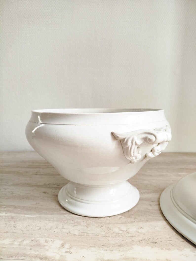 May include: A white ceramic tureen with a smooth, glossy finish, featuring a wide, rounded bowl, a pedestal base, and ornate, scrolled handles on the sides. A matching lid is positioned partially out of frame to the right of the main vessel.