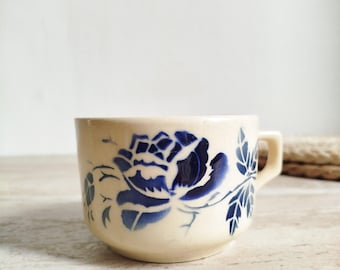 May include: Cream-colored ceramic teacup with a blue floral design. The cup features a large blue flower and leaves, with a small handle. The design is repeated around the cup's exterior.