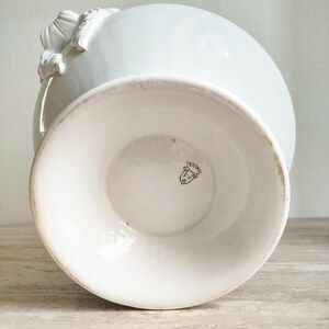 May include: A white ceramic vase with a flared pedestal base and decorative relief detailing on the side. The underside of the base features a small, dark printed maker's mark in the shape of a shield containing the text F.C. and PEXONNE.