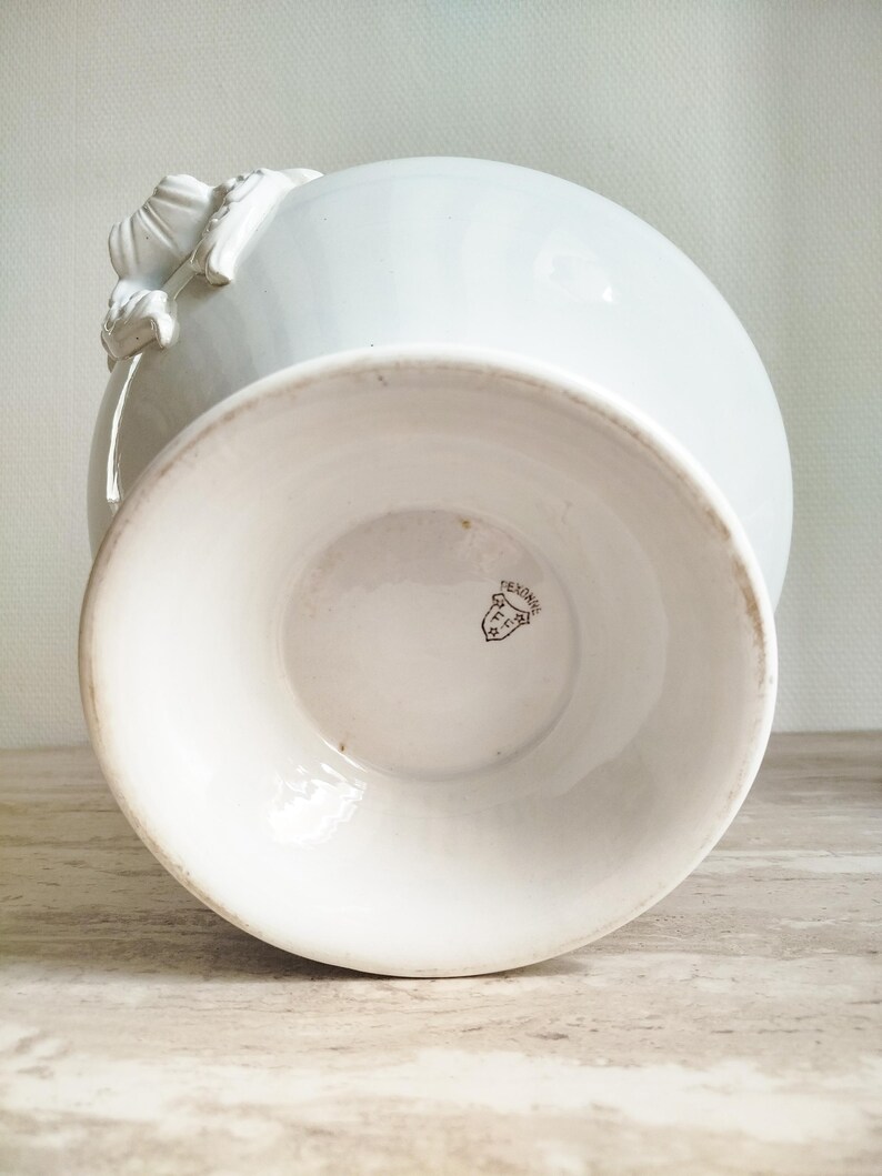 May include: A white ceramic vase with a flared pedestal base and decorative relief detailing on the side. The underside of the base features a small, dark printed maker's mark in the shape of a shield containing the text F.C. and PEXONNE.