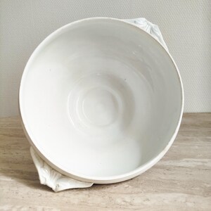 May include: A white ceramic bowl with a smooth, glossy interior and two decorative handles featuring a molded leaf or shell-like pattern on the exterior sides. The bowl has a wide, circular opening and a deep, rounded basin for serving food.
