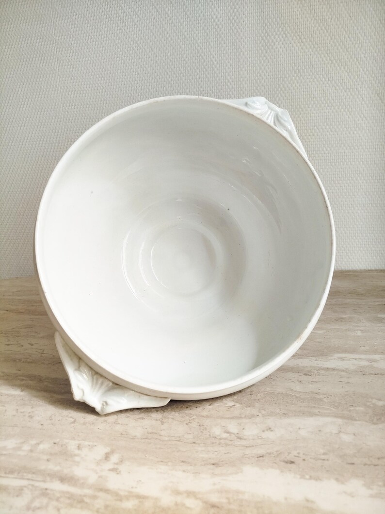 May include: A white ceramic bowl with a smooth, glossy interior and two decorative handles featuring a molded leaf or shell-like pattern on the exterior sides. The bowl has a wide, circular opening and a deep, rounded basin for serving food.