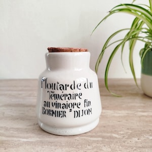 以下が含まれることがあります： コルク栓付きの白いセラミックジャー。ジャーには「Moutarde du Téméraire au vinaigre fin BORNIER DIJON」と黒い文字で書かれています。背景には緑色の植物が見えます。