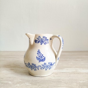 May include: A small, cream-coloured ceramic jug with a blue floral pattern. The jug has a curved handle and a spout. The blue floral design decorates the body and handle of the jug. The jug is likely vintage or antique.