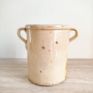 Op de afbeelding: Een cilindrische keramische pot met een glanzend crèmekleurig glazuur en twee handvatten aan de zijkant. Het oppervlak vertoont kleine deukjes en onregelmatigheden over het hele lichaam, met een geel-oranje accent aan de rechteronderkant.