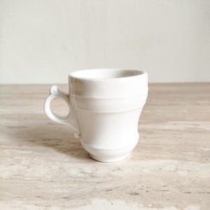 May include: A small, white ceramic teacup with a handle. The cup has a rounded body with horizontal ridges and a small, curved handle. The cup is sitting on a light-colored surface.