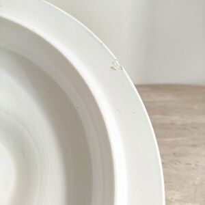 May include: A white ceramic bowl with a smooth, curved rim and a glossy finish. The interior surface shows a subtle, fine crackle pattern throughout the glaze. A small, distinct chip is visible on the outer edge of the rim, revealing the base material.