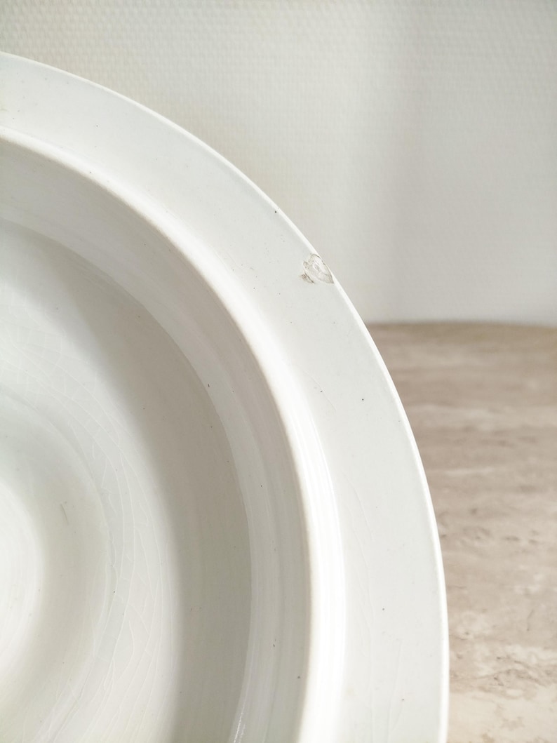 May include: A white ceramic bowl with a smooth, curved rim and a glossy finish. The interior surface shows a subtle, fine crackle pattern throughout the glaze. A small, distinct chip is visible on the outer edge of the rim, revealing the base material.