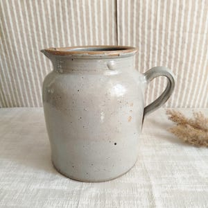 May include: A light gray ceramic pitcher with a speckled finish and a smooth, rounded body. The vessel features a wide, open mouth with a subtle pouring spout, a thick rim, and a sturdy, curved handle attached to the side of the cylindrical base.