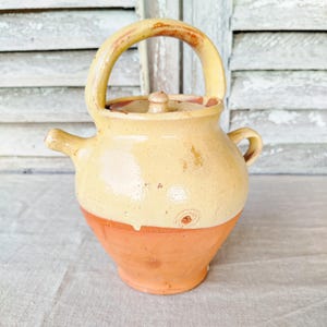 Può includere: Teiera in ceramica rustica fatta a mano, con corpo arrotondato e manico curvo. La teiera è principalmente di colore beige chiaro, con base e dettagli del manico marrone rossiccio. Presenta un piccolo beccuccio e un coperchio.