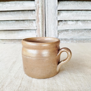 May include: A small, antique-style stoneware mug in a warm, earthy brown hue. The mug features a rounded body with a sturdy handle and subtle horizontal ridges. The rustic design suggests a vintage or farmhouse aesthetic.
