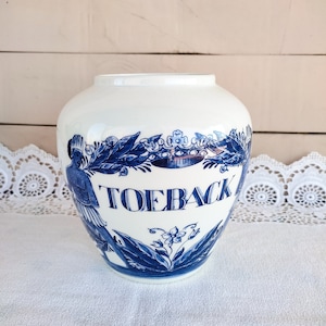 Op de afbeelding: Een witte keramische pot met blauwe en witte bloemenmotieven en het woord "TOEBAC" op de voorkant.