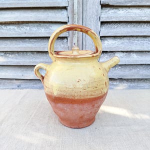 May include: A handmade ceramic teapot with a yellow and terracotta color scheme. The teapot has a rounded body, a curved handle, a spout, and a lid. The teapot is sitting on a neutral-colored surface.