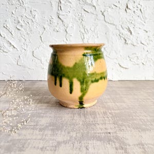 May include: A small, rounded ceramic pot with a tan base colour and a glossy green glaze dripping down from the rim. The vessel features a wide opening, a slightly flared lip, and a narrow base, showcasing a hand-applied drip pattern.