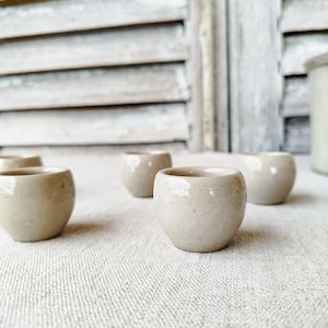 May include: Several small, handmade ceramic cups in a neutral beige hue. The cups have a rounded form and a smooth, glossy finish. They are arranged on a textured, light-coloured surface, suggesting a rustic or minimalist aesthetic.