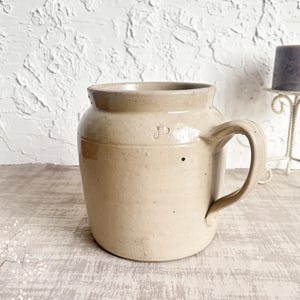 May include: A beige ceramic stoneware mug with a wide, rounded body and a single curved handle. The exterior features a smooth, speckled finish with a small, stamped letter J on the upper side near the rim, showcasing a simple, functional design.