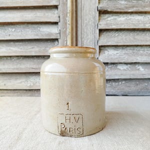 Puede incluir: Un tarro de gres beige, cilíndrico, con tapa redondeada y borde marrón claro. El tarro tiene el número "1" y el texto "H.V. PARIS" estampado en la parte delantera. El fondo presenta una contraventana de madera desgastada.