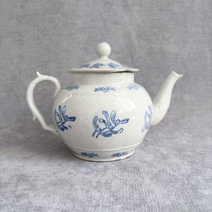 Op de afbeelding: Een witte keramische theepot met een blauw botanisch patroon van maretakken op de buik en het deksel. De theepot heeft een ronde vorm, een gebogen handvat, een korte tuit en een kleine knop in het midden van het deksel om deze te openen.