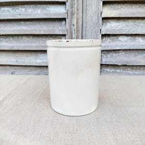 以下が含まれることがあります： わずかに傷んだ縁を持つ、円筒形のオフホワイトのセラミック製の壺。風化した木製のシャッターとニュートラルなトーンのリネンの表面を背景に配置されています。