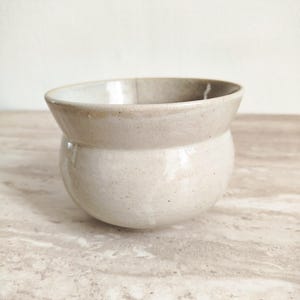 Può includere: Una piccola ciotola in ceramica fatta a mano, con base arrotondata e bordo svasato. La ciotola è di colore bianco sporco macchiettato con un bordo leggermente più scuro. L'interno è beige chiaro. La ciotola è appoggiata su una superficie di marmo.