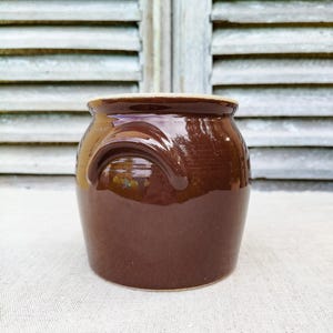 May include: A brown ceramic crock with a glossy finish. The jar has a rounded body, a wide opening, and a handle on the side. The crock is likely used for storage or decoration. The background features a weathered wooden shutter.