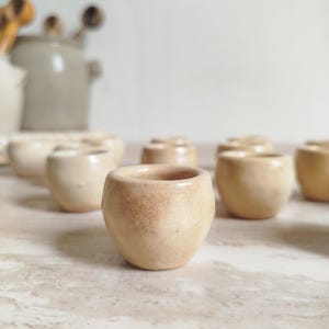 Peut inclure: Ensemble de petites tasses en céramique faites à la main, dans des tons beige et bronzé. Les tasses ont une forme arrondie avec une surface légèrement texturée, suggérant une esthétique rustique ou artisanale. Elles sont disposées sur une surface claire.