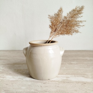 Op de afbeelding: Een beige keramische vaas met twee kleine handvatten en een lichtbruine rand. De vaas bevat gedroogd, veervormig bruin siergras. De vaas staat op een lichtgekleurd, getextureerd oppervlak.