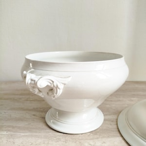 May include: A white porcelain tureen with a smooth, glossy finish, featuring a wide, rounded bowl, a pedestal base, and ornate, sculpted scroll-shaped handles on the sides. A matching white porcelain lid is positioned partially to the right.