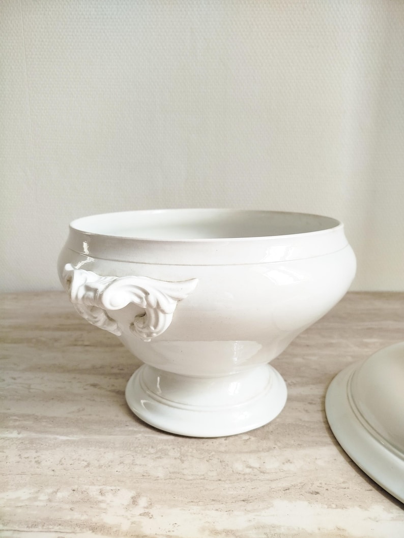 May include: A white porcelain tureen with a smooth, glossy finish, featuring a wide, rounded bowl, a pedestal base, and ornate, sculpted scroll-shaped handles on the sides. A matching white porcelain lid is positioned partially to the right.