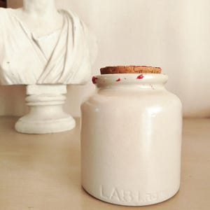 Peut inclure: Un pot en céramique blanche avec un couvercle en liège et des taches rouges. Le pot porte l'inscription "LAB" au fond.