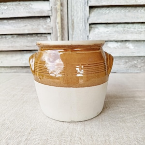 May include: A small, two-toned ceramic pot with a brown upper section and a cream-colored lower section. The brown section features horizontal ridges and small handles on either side. The pot sits on a neutral-colored linen surface.