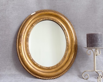 Peut inclure: Un miroir ovale avec un cadre doré présentant un bord extérieur en forme de corde et un motif décoratif intérieur. À côté du miroir se trouve une bougie pilier grise posée sur un bougeoir en métal blanc avec une base à volutes.