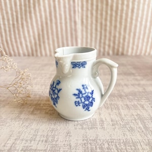 May include: A small white ceramic cream jug featuring a delicate blue floral pattern painted on the exterior. The jug has a curved handle, a small pouring spout, and a rounded body, showcasing a classic design with blue botanical motifs.