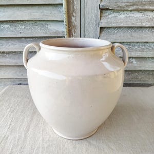 Op de afbeelding: Een crèmekleurige keramische vaas met twee handvatten. De vaas heeft een afgeronde body en een brede opening. De achtergrond toont verweerde houten luiken en een neutraal gekleurde linnen doek.