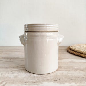 Puede incluir: Un tarro de cerámica beige con tapa. El tarro tiene dos asas pequeñas y estrías horizontales alrededor del centro y en la tapa. Está sobre una superficie clara. Un posavasos tejido está en el fondo.