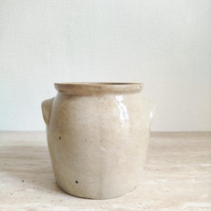 May include: A beige ceramic jar with a wide opening and two small handles. The jar has a slightly rounded shape and a light brown rim. The surface has a speckled appearance, and the jar is set against a neutral background.