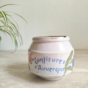 Op de afbeelding: Een kleine, gebroken witte keramische pot met een licht uitlopende basis en een afgeronde bovenkant. De pot heeft de tekst "Confitures d'Auvergne" in blauw schrift, met bloemaccenten in roze en blauw. De pot is waarschijnlijk vintage.