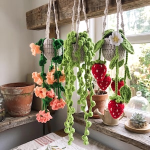 May include: Three crocheted hanging planters suspended from a wooden beam. The left features peach-colored flowers, the center a green string of pearls plant, and the right red strawberries with a white flower, all in light gray pots.