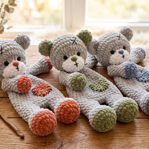 May include: Three handmade crochet teddy bears made from soft grey yarn. Each bear features a white muzzle, black button eyes, and a stitched mouth. The bears have contrasting coloured accents on their ears, hands, and feet in orange, green, and blue.