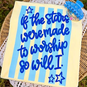 May include: A light yellow t-shirt with blue vertical stripes and the text "if the stars were made to worship so will I." A blue hair clip and bracelets are on the shirt.